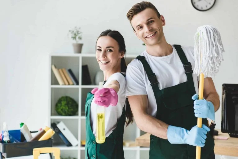 happy-young-cleaners-with-mop-and-spray-bottle-smiling-at-camera-while-cleaning-office-768x513